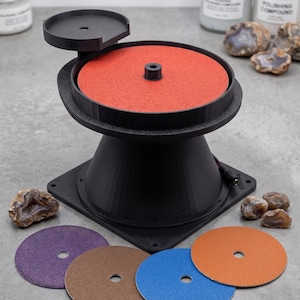 May include: A black rotary lapidary machine with a central red abrasive disc, a small side platform, and four additional circular abrasive discs in purple, brown, blue, and orange. The device features a conical base and a central mounting spindle.