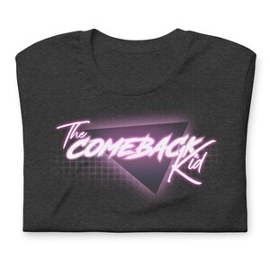 May include: A dark grey t-shirt with a pink neon graphic design that says "The Comeback Kid" in a retro 80s style font. The graphic features a triangle with a grid pattern behind it.