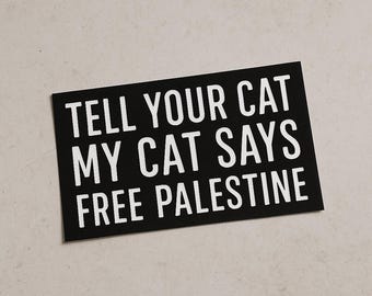 May include: A black rectangular sticker with white text. The text reads "TELL YOUR CAT MY CAT SAYS FREE PALESTINE". The sticker is on a light beige surface.