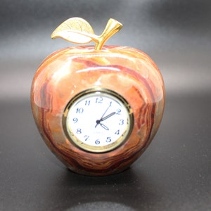 May include: A red and brown marble apple clock with gold accents and a leaf on top. The clock face has black hands and white numbers.