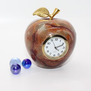 May include: A brown and white marble apple clock with a gold leaf and stem. The clock face is white with black numbers and hands. The clock is sitting on a white surface with three blue marbles.