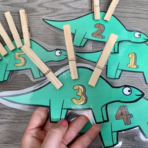 Op de afbeelding: Vijf groene, dinosaurusvormige kaarten met de nummers 1 tot en met 5 erop gedrukt. Elke kaart heeft een houten wasknijper eraan bevestigd. De wasknijpers worden gebruikt om het aantal objecten te tellen.