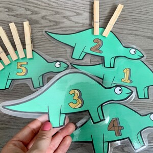 May include: Five green dinosaur-shaped cards with numbers 1 through 5 printed on them. Each card has a space for a clothespin on the back. There are clothespins on the cards with numbers 1, 2, and 5.