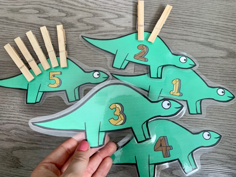 May include: Five green dinosaur-shaped cards with numbers 1 through 5 printed on them. Each card has a space for a clothespin on the back. There are clothespins on the cards with numbers 1, 2, and 5.