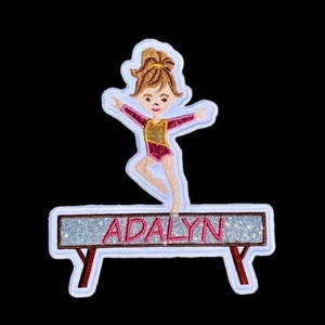 Pode incluir: Um remendo branco, brilhante, bordado de uma ginasta em uma trave de equilíbrio com o nome "ADALYN" na trave.
