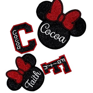 Pode incluir: Dois apliques de cabeça de Mickey Mouse preto e vermelho com glitter com os nomes "Cocoa" e "Faith" escritos em branco. Uma letra "C" preta e vermelha com glitter e uma letra "F" preta e vermelha com glitter com o texto "Faith" escrito em branco.