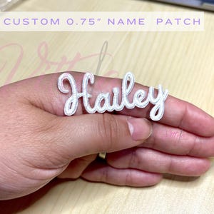 May include: White embroidered name patch with the name "Hailey" in a cursive font. The patch is 0.75 inches tall.