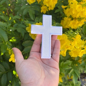 Op de afbeelding: Witte vilt kruis patch met een witte omtrek. De patch wordt in een hand gehouden tegen een achtergrond van gele bloemen en groene bladeren.