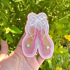 Puede incluir: Un parche de zapato de ballet de purpurina rosa y dorada con una cinta blanca. El parche está bordado con una pequeña flor verde en cada zapato.
