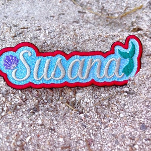May include: A blue and red embroidered patch with the name "Susana" in silver glitter lettering. A purple seashell and a green mermaid tail are on either side of the name.