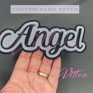 Puede incluir: Un parche de nombre personalizado con el nombre "Angel" en un diseño negro, plateado brillante y blanco. El parche tiene un borde negro y se muestra sobre un fondo gris oscuro. El texto "CUSTOM NAME PATCH" está en la parte superior.