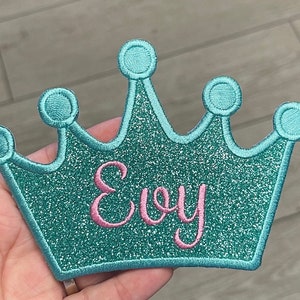 Puede incluir: Un parche de corona de purpurina turquesa con el nombre "Evy" bordado en rosa. La corona tiene un contorno bordado detallado y detalles circulares. Este parche es adecuado para ropa o accesorios.
