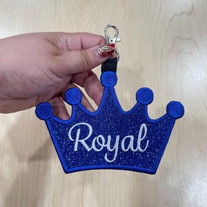 May include: A blue crown-shaped keychain with the word "Royal" embroidered in white script. The crown has a glittery texture and is attached to a silver-colored clasp and a black strap. The keychain is designed for decorative purposes.