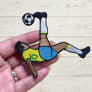 Puede incluir: Parche bordado con un futbolista con uniforme amarillo y azul pateando un balón. El parche tiene detalles en negro, marrón, blanco y verde. El jugador está en pleno movimiento de patada.