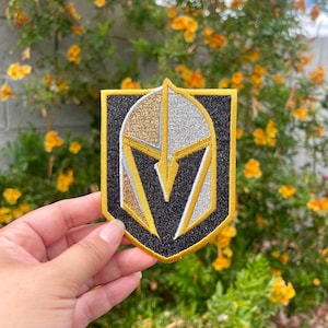May include: A black, gold, and silver embroidered patch with a knight's helmet design. The helmet is outlined in gold and has a black and white V shape in the center.