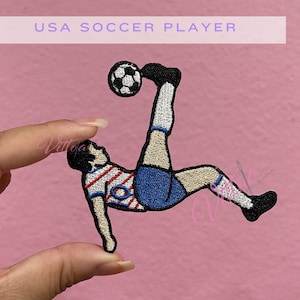 May include: An embroidered patch featuring a soccer player performing a bicycle kick with a black and white soccer ball. The player wears a red and white striped shirt, blue shorts, white socks, and black shoes. The text USA SOCCER PLAYER is visible.