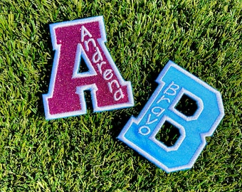 May include: A glittery dark pink letter A patch with the name Andrena embroidered in white, and a light blue letter B patch with the name Bravo embroidered in white. Both varsity-style patches feature a thick white border and a stitched edge.