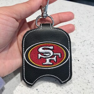 Könnte beinhalten: Schwarzer Leder-Handdesinfektionsmittelhalter mit weißem Stepprand und einem gestickten San Francisco 49ers-Logo in Rot, Weiß und Gold. Der Halter hat einen silbernen Karabinerhaken.