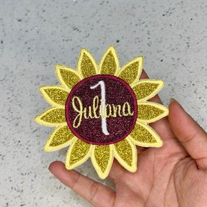 Puede incluir: Un girasol de purpurina dorada con un centro burdeos. El centro tiene el número "1" y el nombre "Juliana" escrito en blanco.