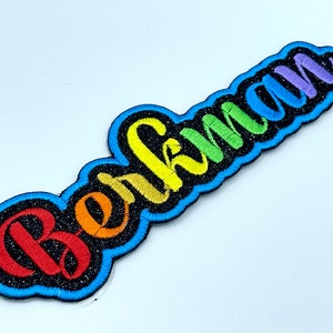 May include: Rainbow colored embroidered patch with the word "Bekman" in a cursive font. The patch has a black background and a blue outline.