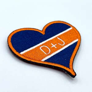 Op de afbeelding: Een hartvormige patch met een donkerblauwe achtergrond en een oranje streep in het midden. De streep heeft witte tekst die "D+J" luidt.