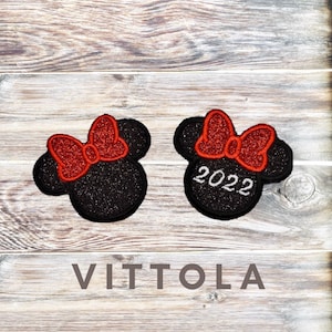 Pode incluir: Dois ornamentos de cabeça de rato pretos com glitter e laços vermelhos com glitter. O ornamento à direita tem o ano 2022 escrito em glitter branco. Os ornamentos estão sobre um fundo de madeira com a palavra "VITTOLA" escrita abaixo.
