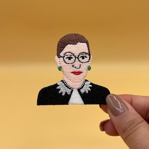 Puede incluir: Parche bordado para planchar con un retrato de Ruth Bader Ginsburg en su toga judicial. Lleva gafas y tiene una sonrisa cálida.