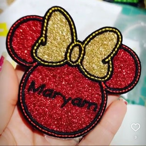 Pode incluir: Patch de ferro vermelho e dourado com glitter, com um laço e o nome "Maryam" em bordado preto.