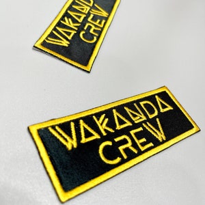 Op de afbeelding: Twee zwarte stoffen patches met gele geborduurde tekst die "Wakanda Crew" luidt.