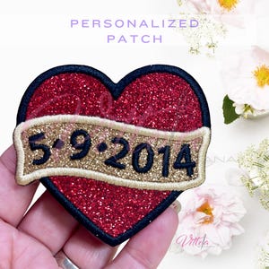 May include: A heart-shaped patch with red glitter and a black border. A gold banner across the heart displays the date "5.9.2014" in black. The patch is labeled "PERSONALIZED PATCH" in purple text.