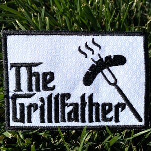 Może przedstawiać: Haftowana naszywka z napisem "The Grillfather" w kolorze czarnym, z grafiką kiełbasy na widelcu z dymem. Naszywka ma białe tło z rombowym wzorem i czarną obwódką.