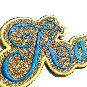 Könnte beinhalten: Nahaufnahme eines goldglitzernden Aufnähers mit dem Namen "Kay" in blauer Schrift gestickt. Der Aufnäher hat einen gelben Rand und eine schwarze Rückseite, geeignet für Kleidung oder Accessoires.