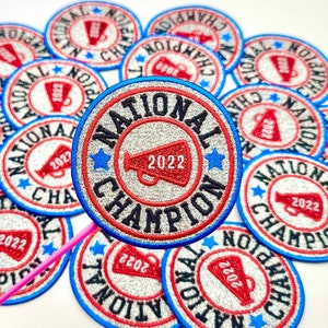 May include: A close-up of a silver glitter patch with a red and blue border. The patch says "National Champion 2022" with a red megaphone graphic in the center.
