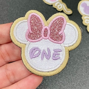 May include: A white and gold embroidered patch with a pink glitter bow and the word "ONE" in purple. The patch is shaped like a mouse head.
