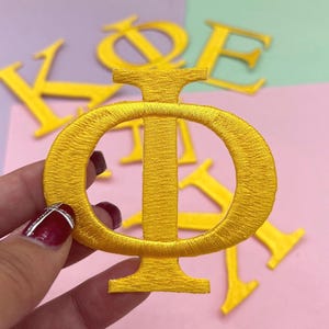 May include: A close-up of a gold embroidered Greek letter Phi patch, held by a hand. Other gold embroidered Greek letters are scattered in the background. The patches are on a pink, green, and blue background.