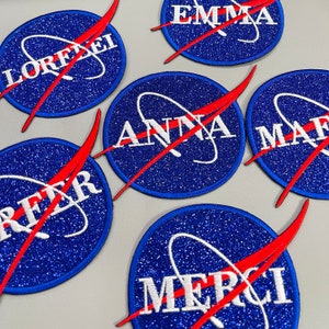 May include: Six blue and white embroidered patches with red stitching. The patches feature the words "Emma", "Lorelei", "Anna", "MaF", "RFR", and "Merci" in white lettering. The patches are in the shape of a circle with a red and white design resembling a rocket ship.