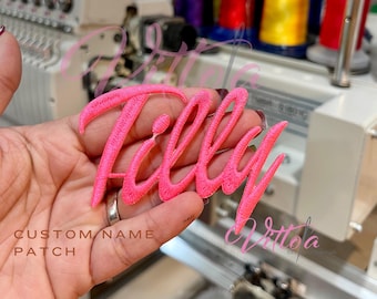 May include: A pink embroidered patch with the name "Tilly" in a cursive font. The patch is held in a hand and is ready to be ironed onto a garment.