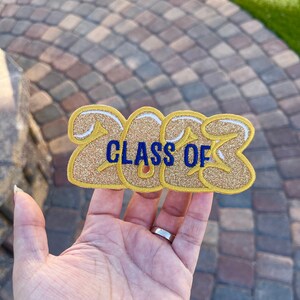 May include: Gold glitter iron-on patch with the text "CLASS OF 2023" in blue thread.