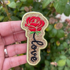 May include: A gold glitter iron-on patch with a red embroidered rose and the word "love" written in black cursive.