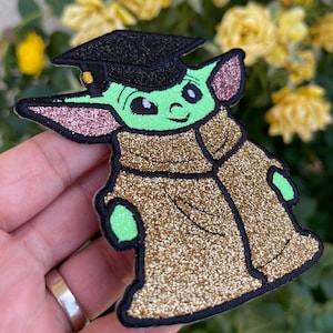 May include: A green glitter patch of a character wearing a black graduation cap and a gold glitter robe. The character has large ears and a friendly expression.