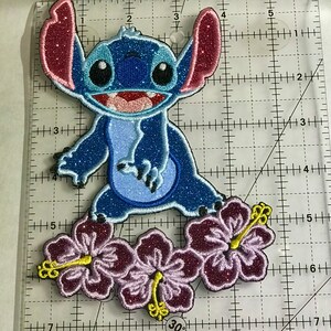 Op de afbeelding: Een blauwe en roze glinsterende geborduurde patch van Stitch uit Lilo & Stitch, staand op drie paarse hibiscusbloemen.