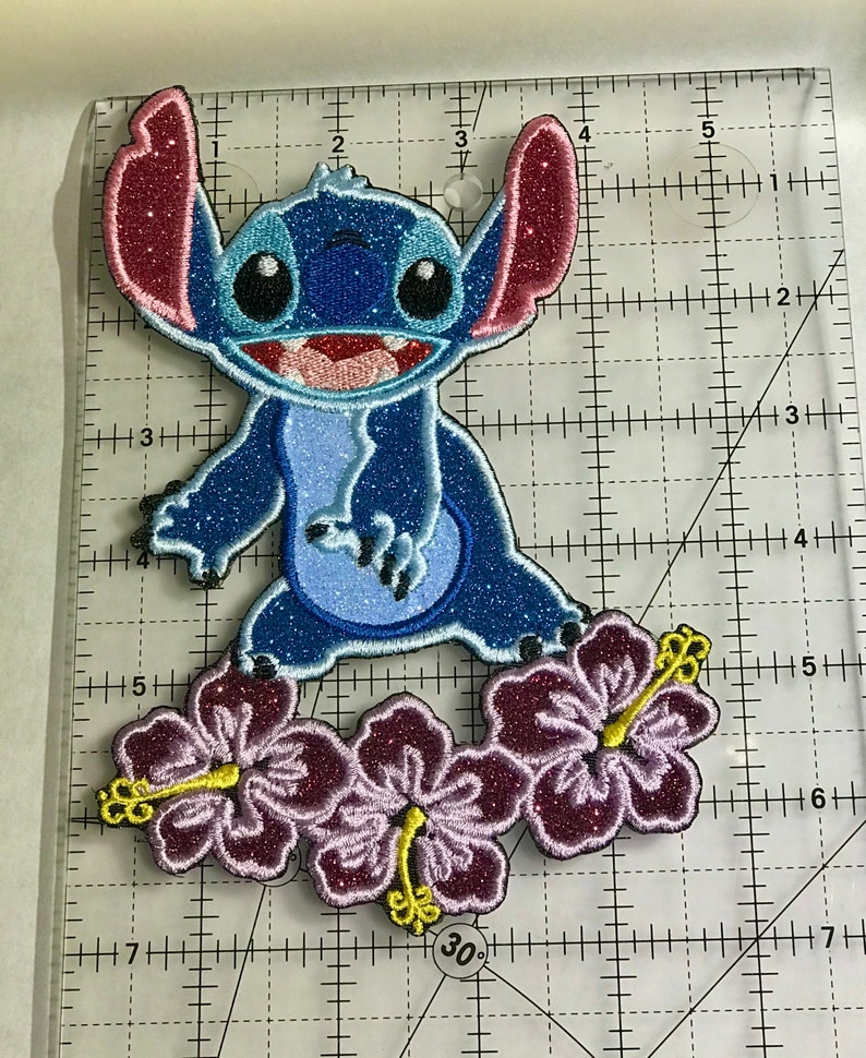 Op de afbeelding: Een blauwe en roze glinsterende geborduurde patch van Stitch uit Lilo & Stitch, staand op drie paarse hibiscusbloemen.