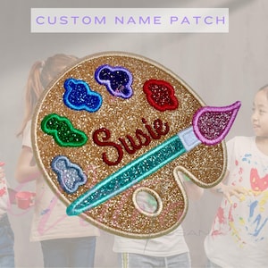 May include: A glittery gold custom name patch in the shape of an artist's palette. The patch features colorful paint splotches, a paintbrush, and the name "Susie" embroidered in red. The text "CUSTOM NAME PATCH" is at the top.