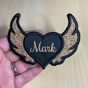 Può includere: Toppa a forma di cuore nera con ali dorate con glitter e il nome "Mark" ricamato in filo dorato.