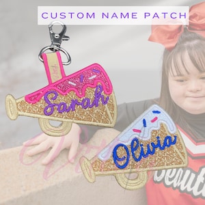 May include: Custom name patches in the shape of a megaphone. One is pink and gold with the name "Sarah" embroidered in purple. The other is blue and gold with the name "Olivia" embroidered in blue. Both have a clip for easy attachment.