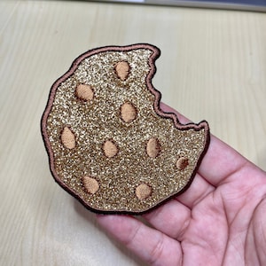 Könnte beinhalten: Ein brauner Glitzerkeks mit einem herausgebissenen Stück. Der Keks hat einen braunen Rand und ist mit braunen Schokoladenstückchen verziert.