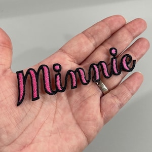May include: Pink felt iron-on patch with the word "Minnie" in a cursive font with black outlining.