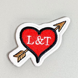May include: A white felt patch with a red heart pierced by a gold arrow. The heart has a black outline and the letters "L&T" in white inside.