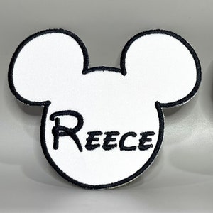 Pode incluir: Cabeça de Mickey Mouse de feltro branco com contorno preto e o nome Reece bordado em preto.