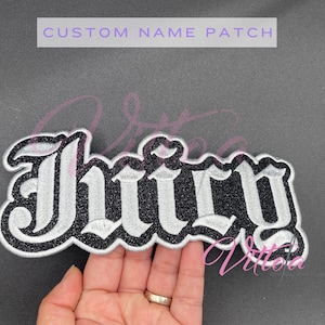 May include: A custom name patch with the word "Juicy" in a gothic font. The patch has a black glitter background with white embroidered lettering and a black border. The text "CUSTOM NAME PATCH" is above the patch.